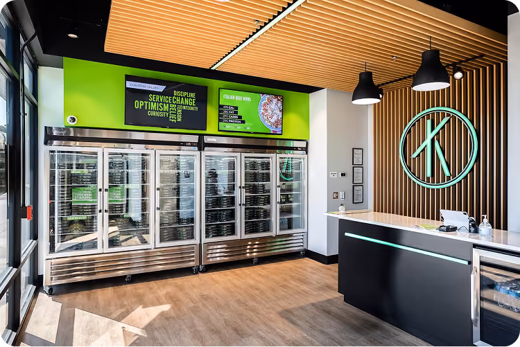 Modern kitchen with glass-door fridges, wood floor, wooden ceiling slats, and black counter lit by pendant lamps.
