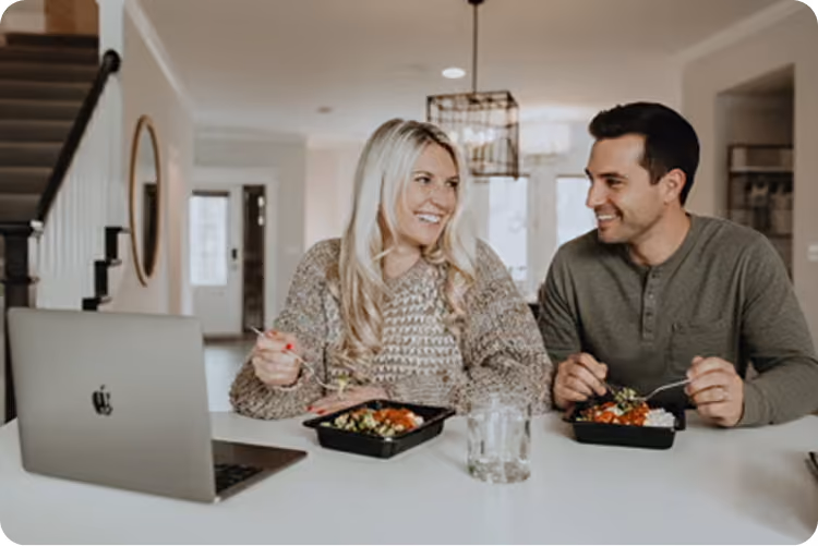 A smiling couple eating meals at a table while looking at a laptop in a modern kitchen.
