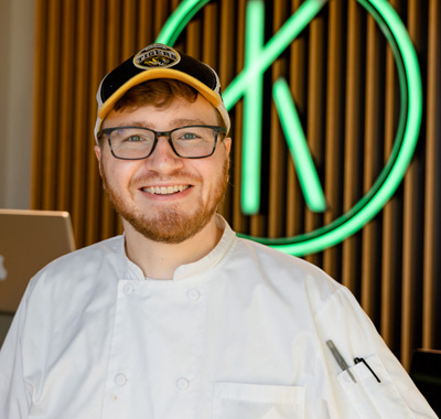 Smiling chef with glasses, white coat, and cap in front of wooden wall with green neon logo.
