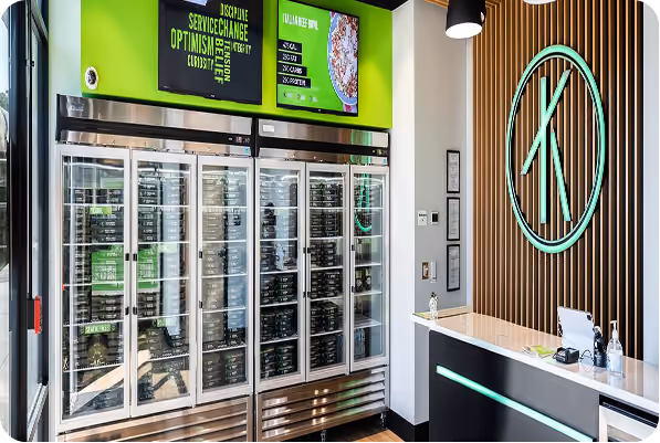 Modern store interior with refrigerated shelves and a counter under a green and wood-paneled wall with a large logo.