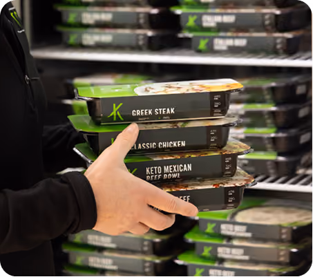 Person holding stacked packaged ready meals labeled Greek Steak, Classic Chicken, Keto Mexican Beef Bowl.