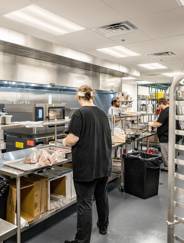 Three people preparing food in a clean, industrial kitchen with stainless steel counters and equipment.