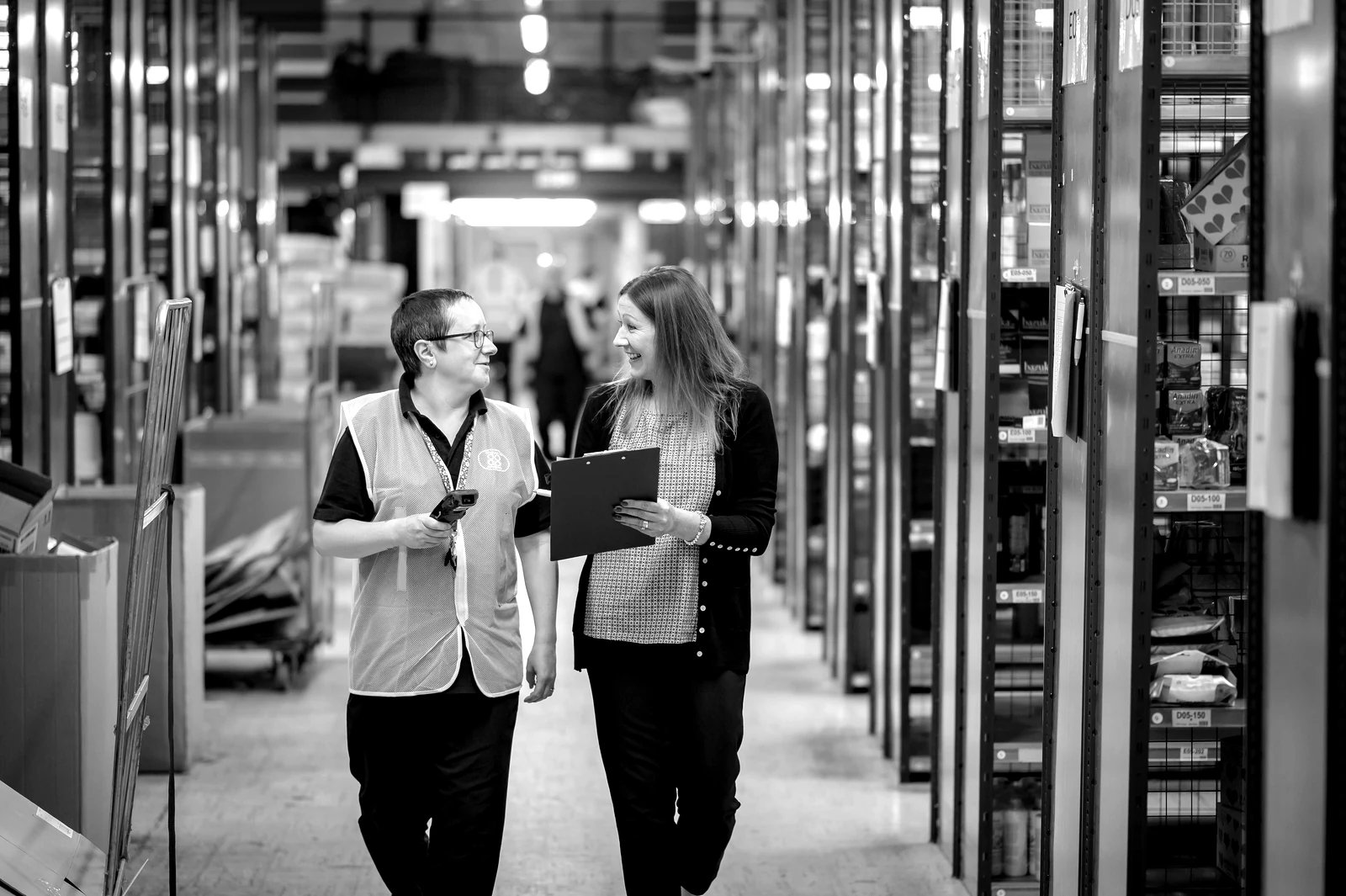 Two warehouse staff reviewing a clipboard as they walk an aisle