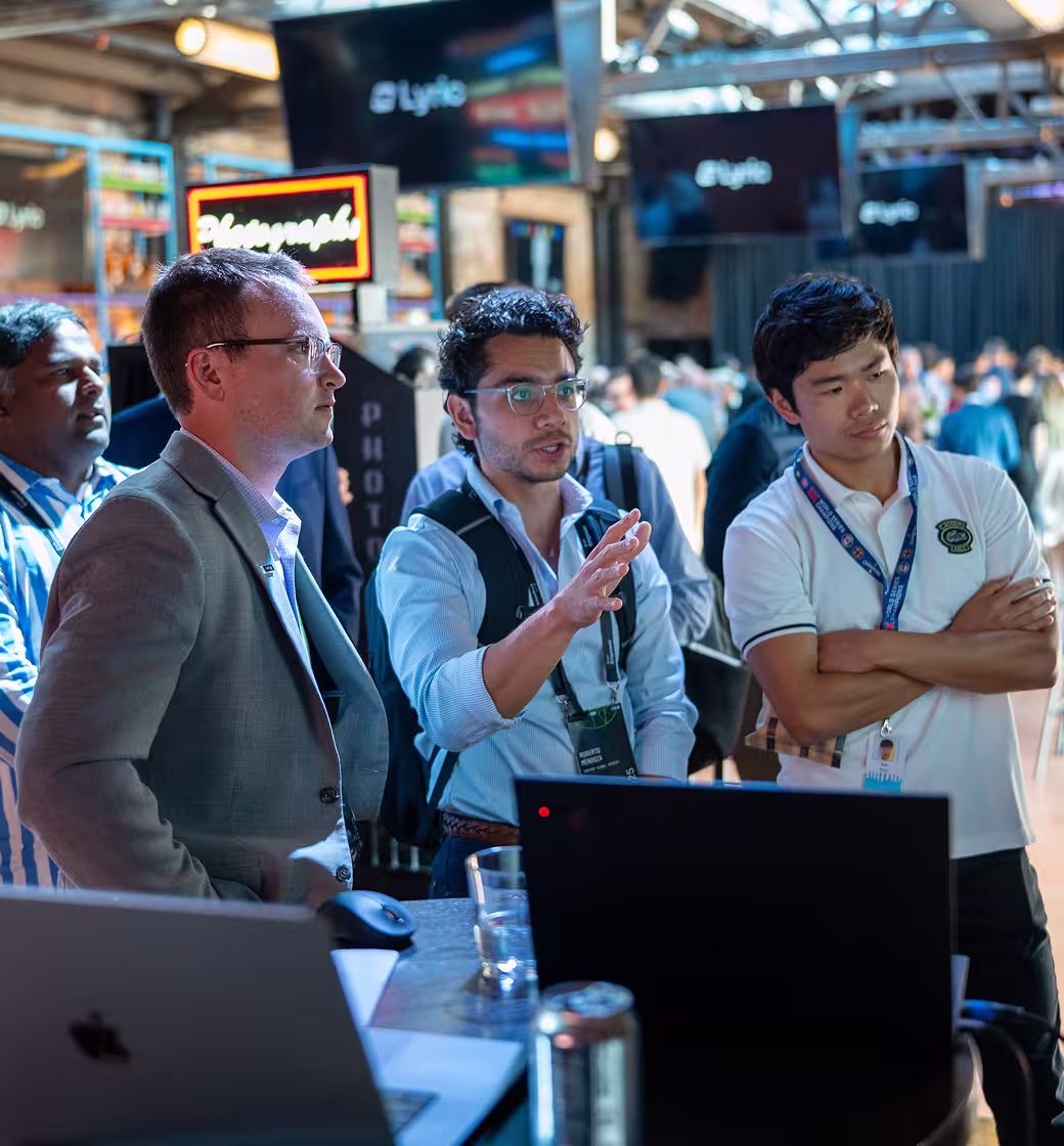 Four men at a lyric event, standing and engaged in discussion with laptops, with a busy conference background.