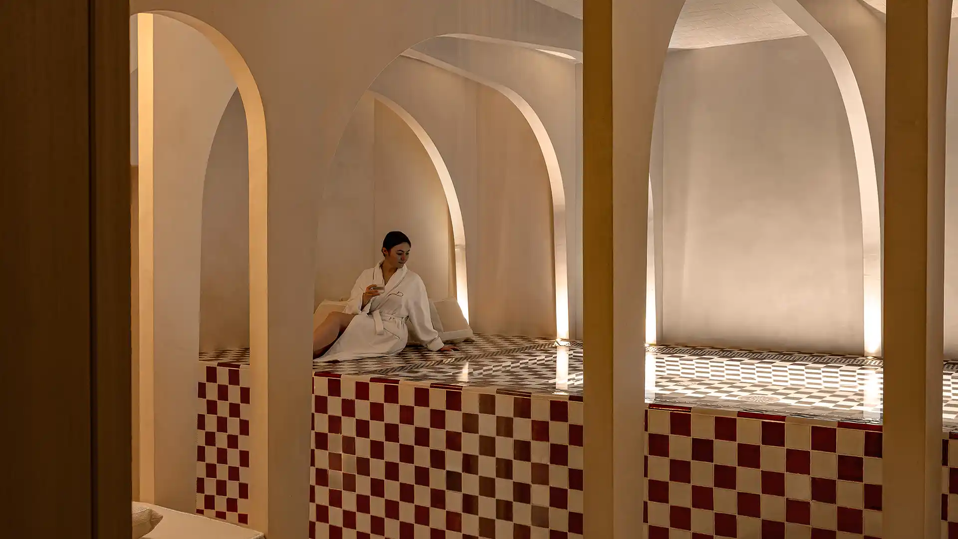 Femme en peignoir blanc assise dans le hammam voûté au carrelage à damier rouge et blanc spa Château des Fleurs