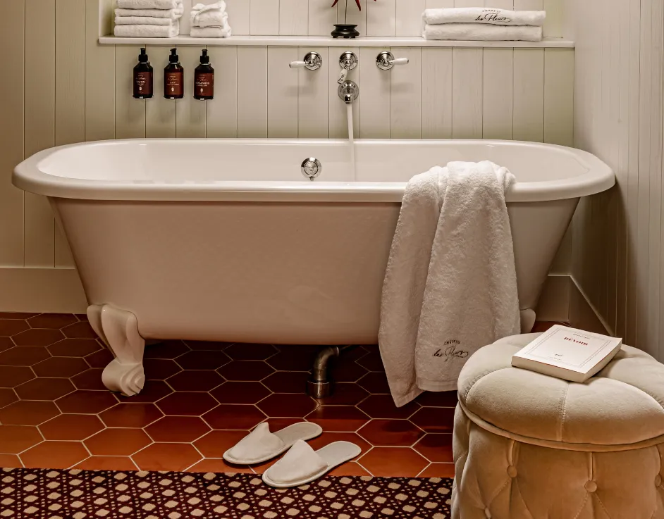 Baignoire sur pieds avec serviettes brodées chaussons et pouf sur carrelage terracotta Château des Fleurs