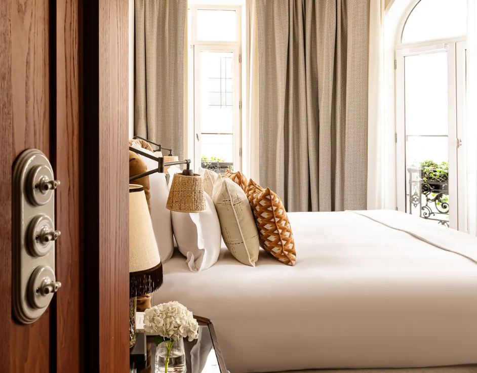 Vue depuis l'entrée de la chambre avec lit coussins motif géométrique et grande fenêtre sur balcon fleuri Château des Fleurs