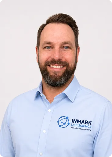 Smiling man with beard wearing a light blue Inmark Life Science shirt against a plain background.