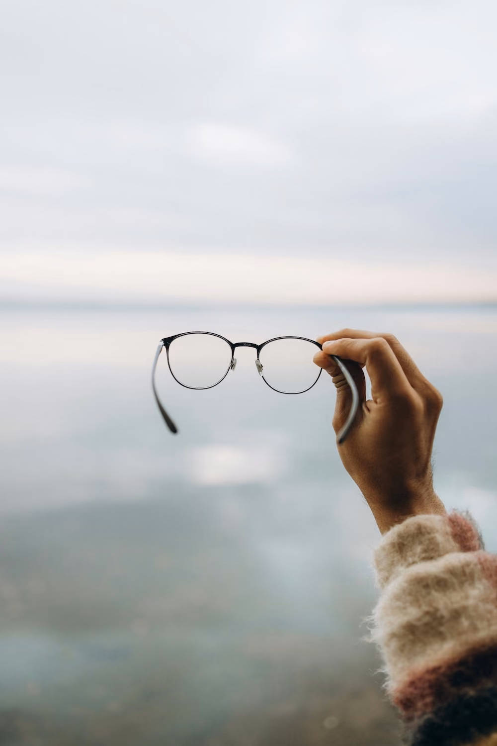 A hand wearing glasses is holding them up near a body of water with trees in the background.