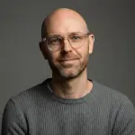 Bald man with glasses and beard wearing a gray ribbed sweater against a dark background.
