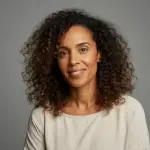 Smiling woman with curly hair wearing a white top against a plain gray background.