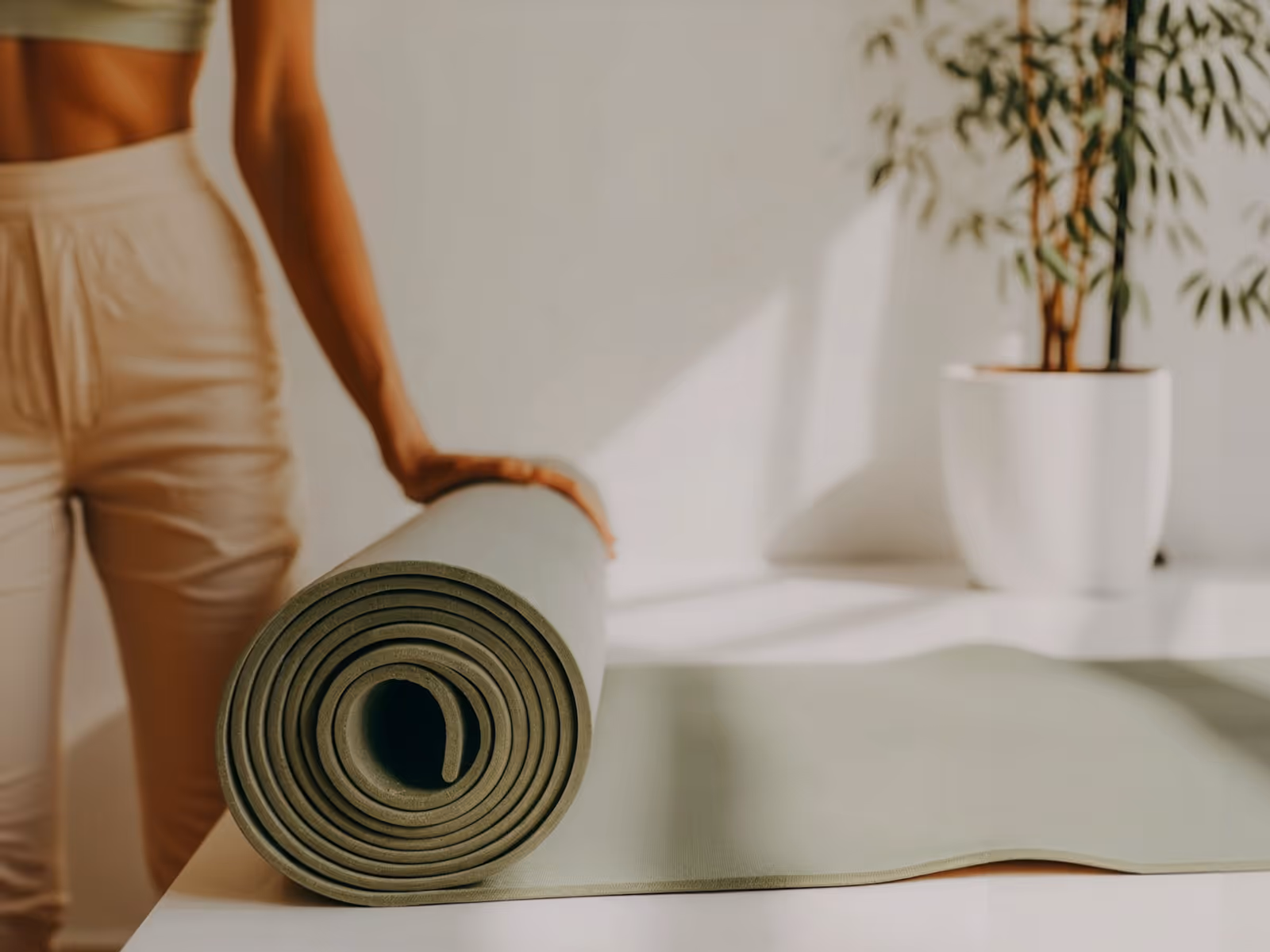 Close-up of a rolled green yoga mat on a white surface with a person in beige pants holding it.