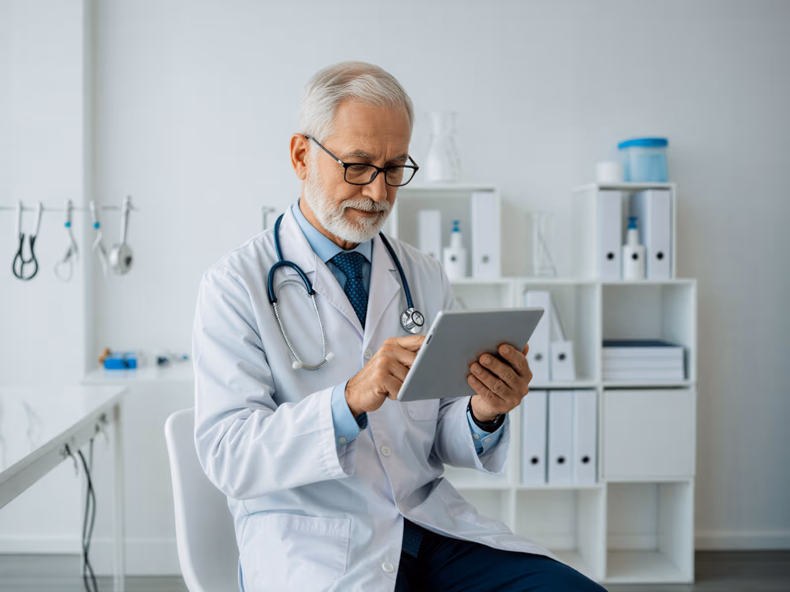 Senior male doctor in white coat using a tablet in a modern medical office.