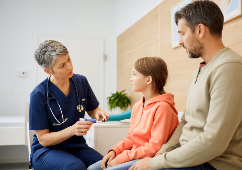 Female doctor in scrubs talking to a girl in a pink hoodie with a man, likely her father, in a clinic.