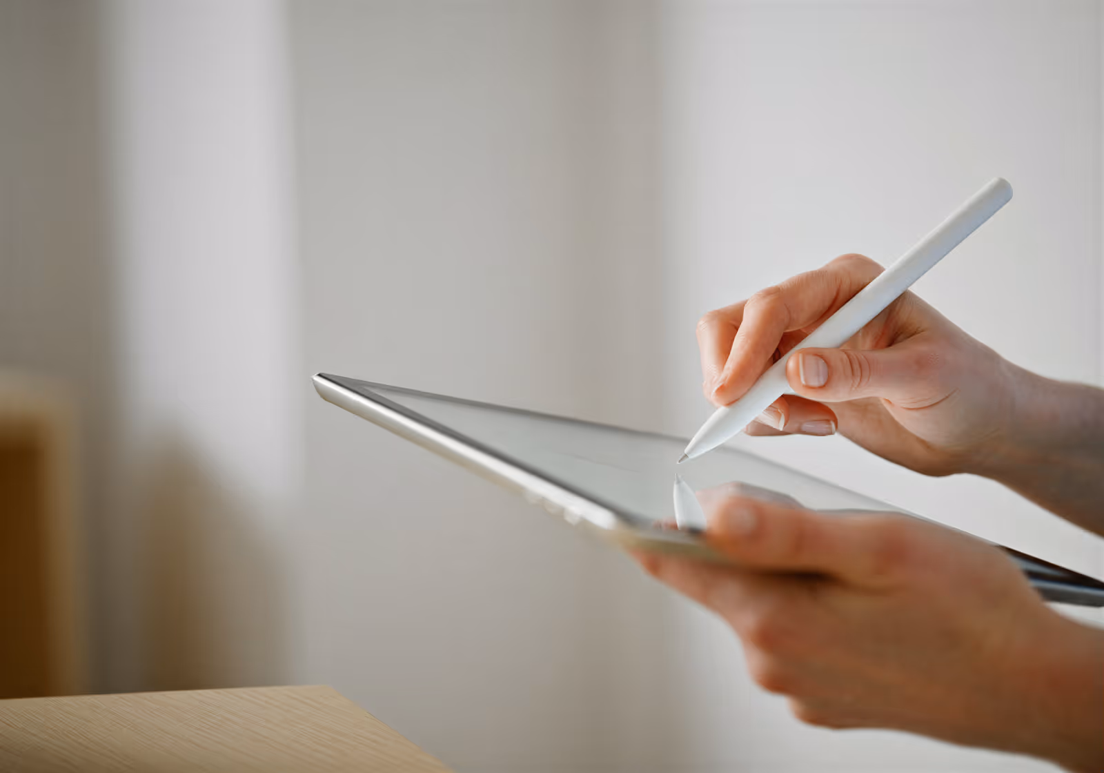 Hands holding a tablet and using a white stylus pen on its screen in a neutral setting.
