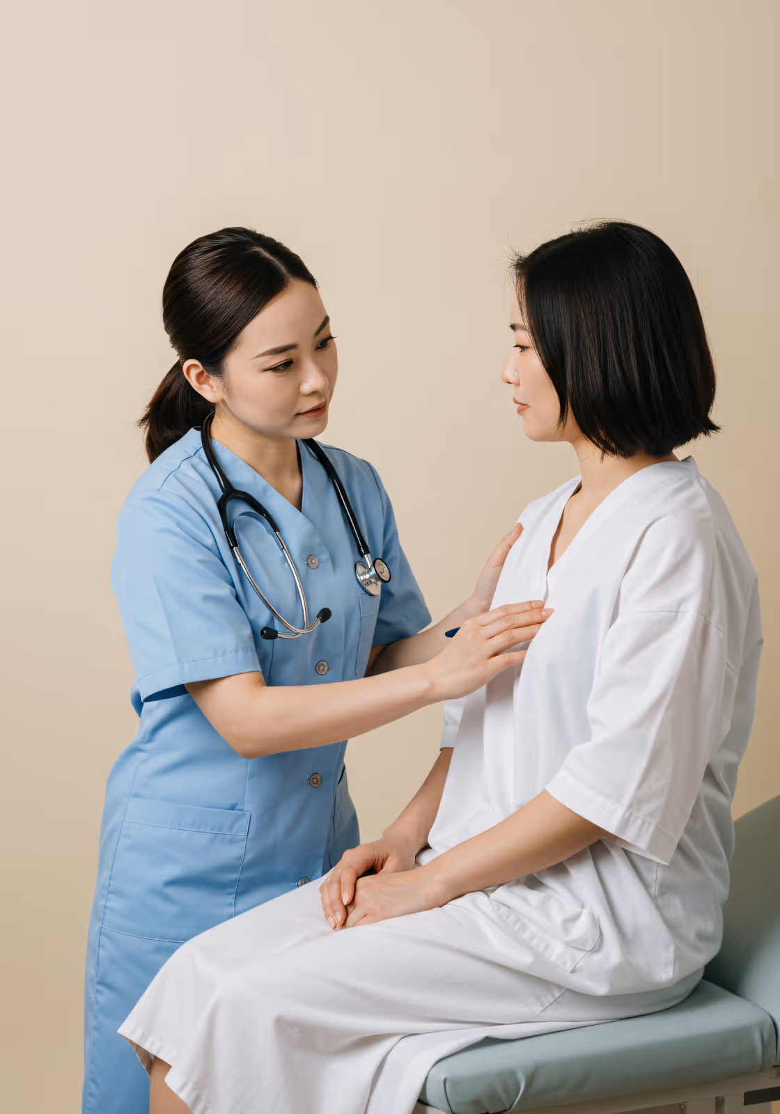 Female doctor in blue scrubs examines a seated female patient wearing a white gown.