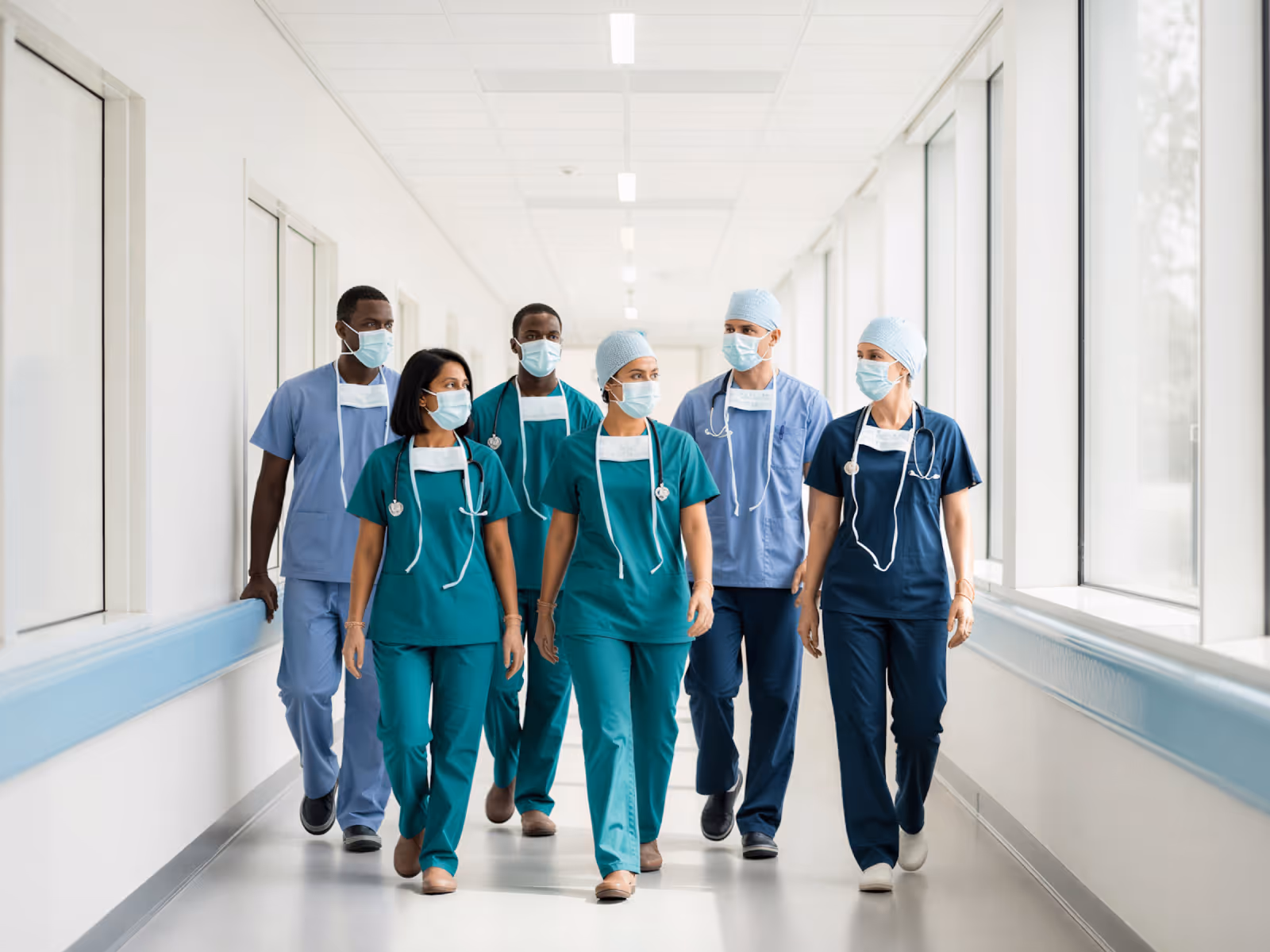 Group of six masked healthcare professionals walking down a bright hospital corridor.