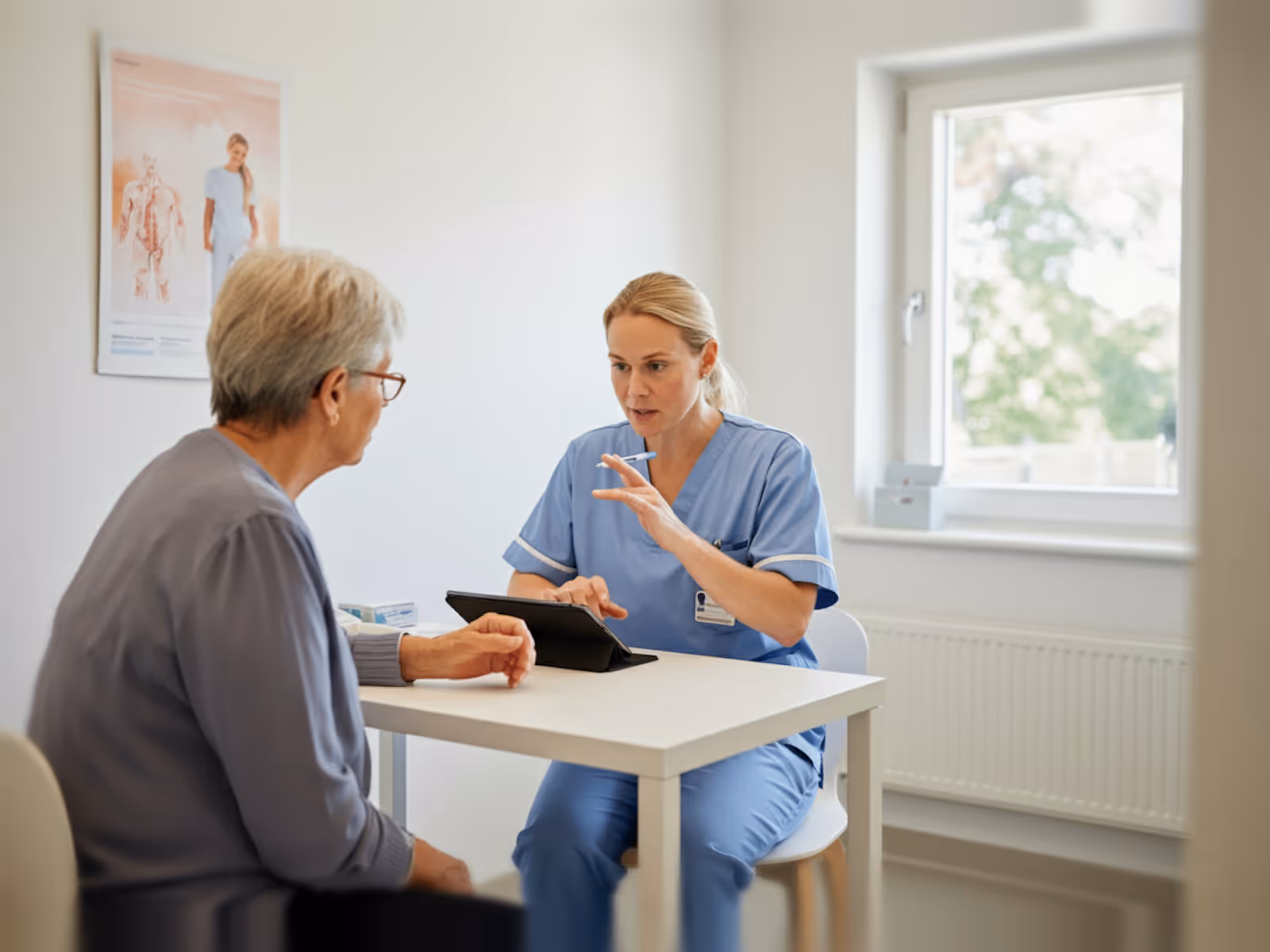 Nurse in blue scrubs consulting with an elderly woman at a white table in a bright room.