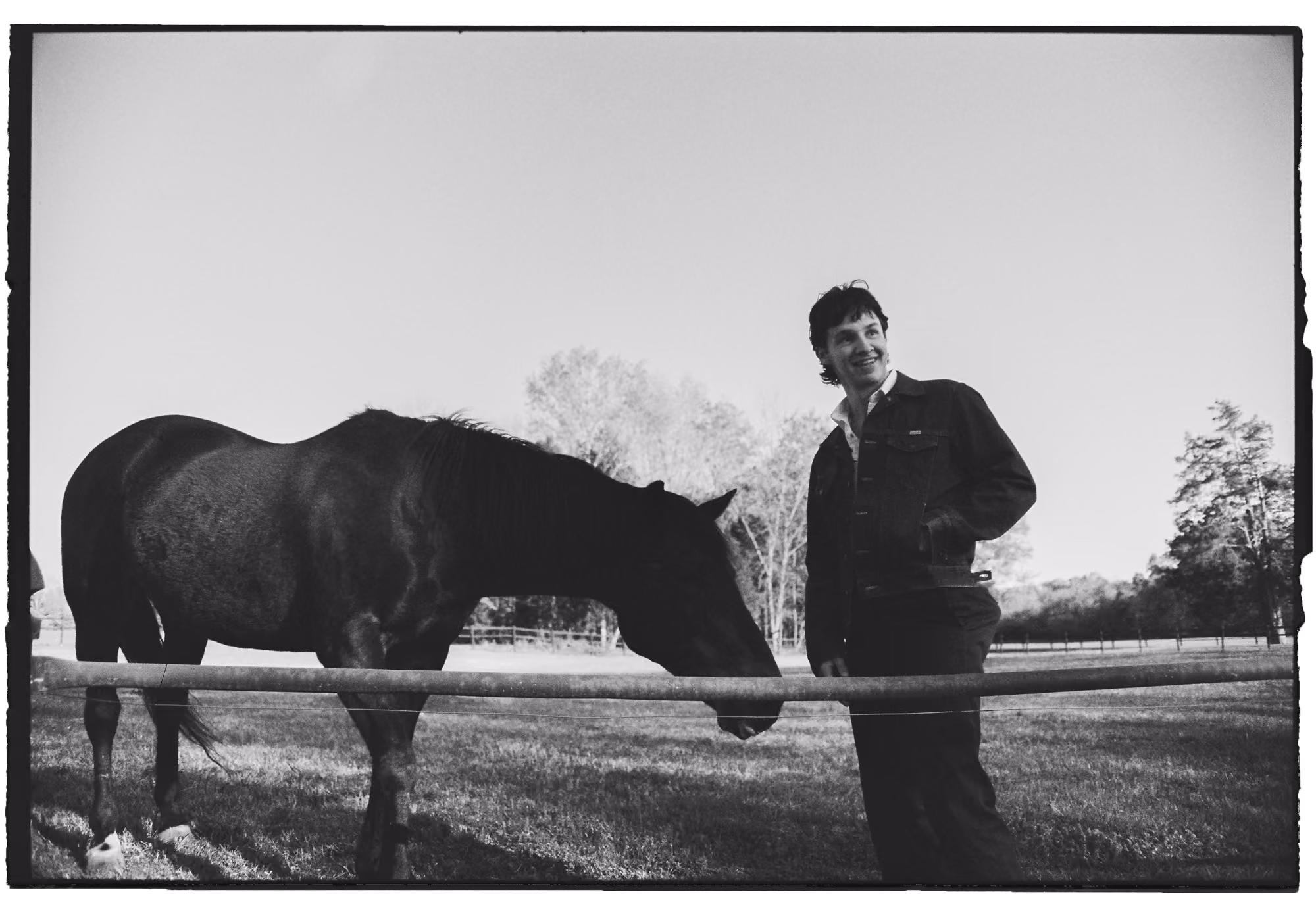 Jack Blocker standing near a black horse behind a fence in a grassy field with trees in the background.
