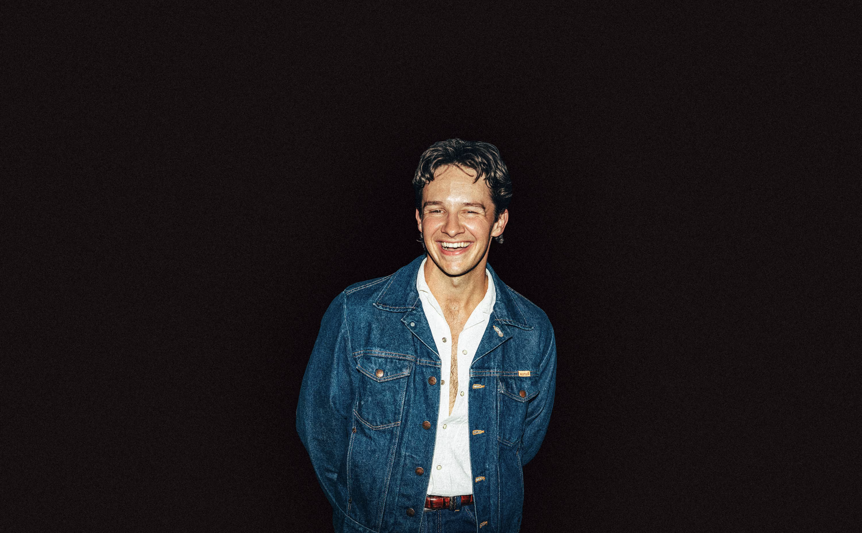 Jack Blocker a Young man smiling in a denim jacket and white shirt against a black background.
