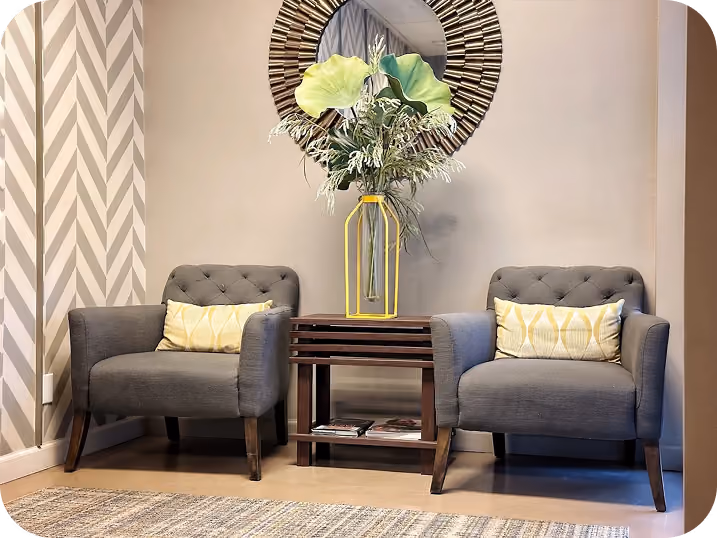 Two gray armchairs with yellow patterned pillows beside a wooden table holding a tall vase with green leaves.