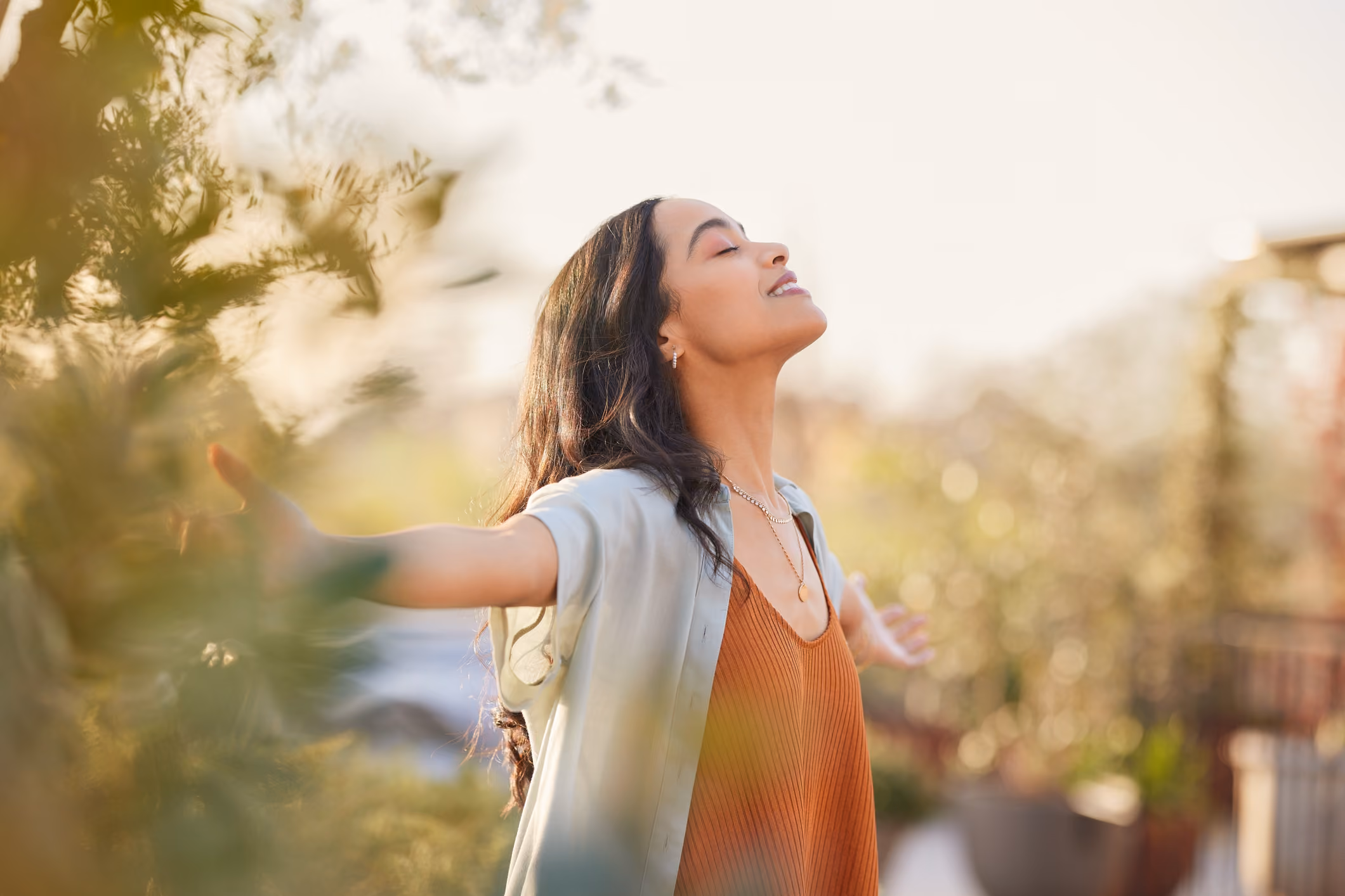 Woman standing outdoors with eyes closed and arms outstretched enjoying sunlight.