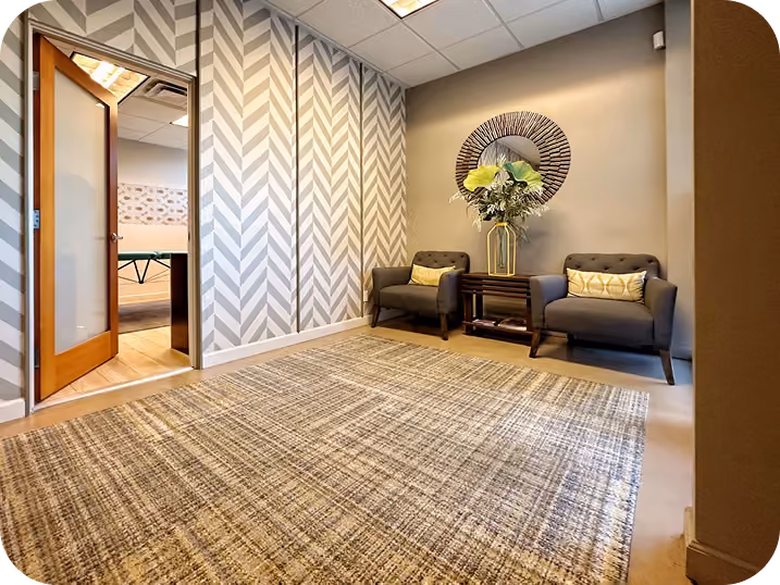 Modern waiting area with two dark chairs, a small table with flowers, round mirror, and textured wall and carpet.