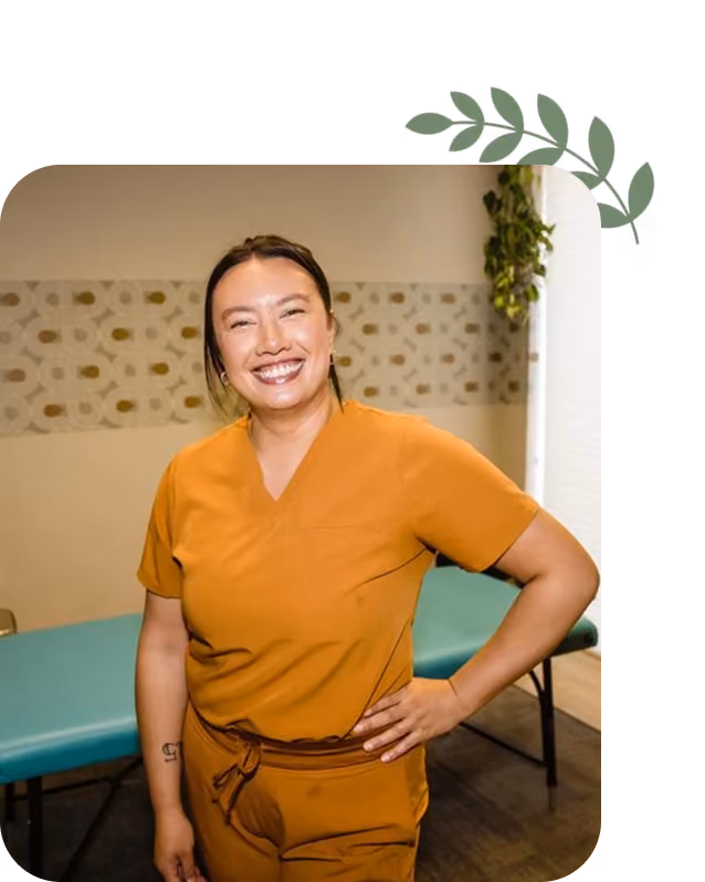 Smiling woman in orange scrub uniform stands with hand on hip in a medical exam room.