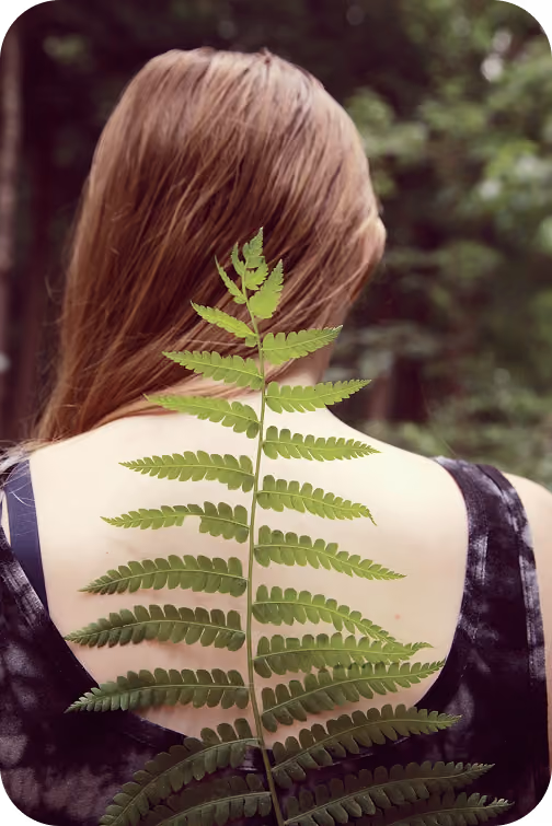 Person with long hair seen from the back holding a green fern leaf over their bare upper back.