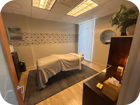 Massage room with a covered massage table, soft lighting, a plant, and tissue box on dark wood furniture.