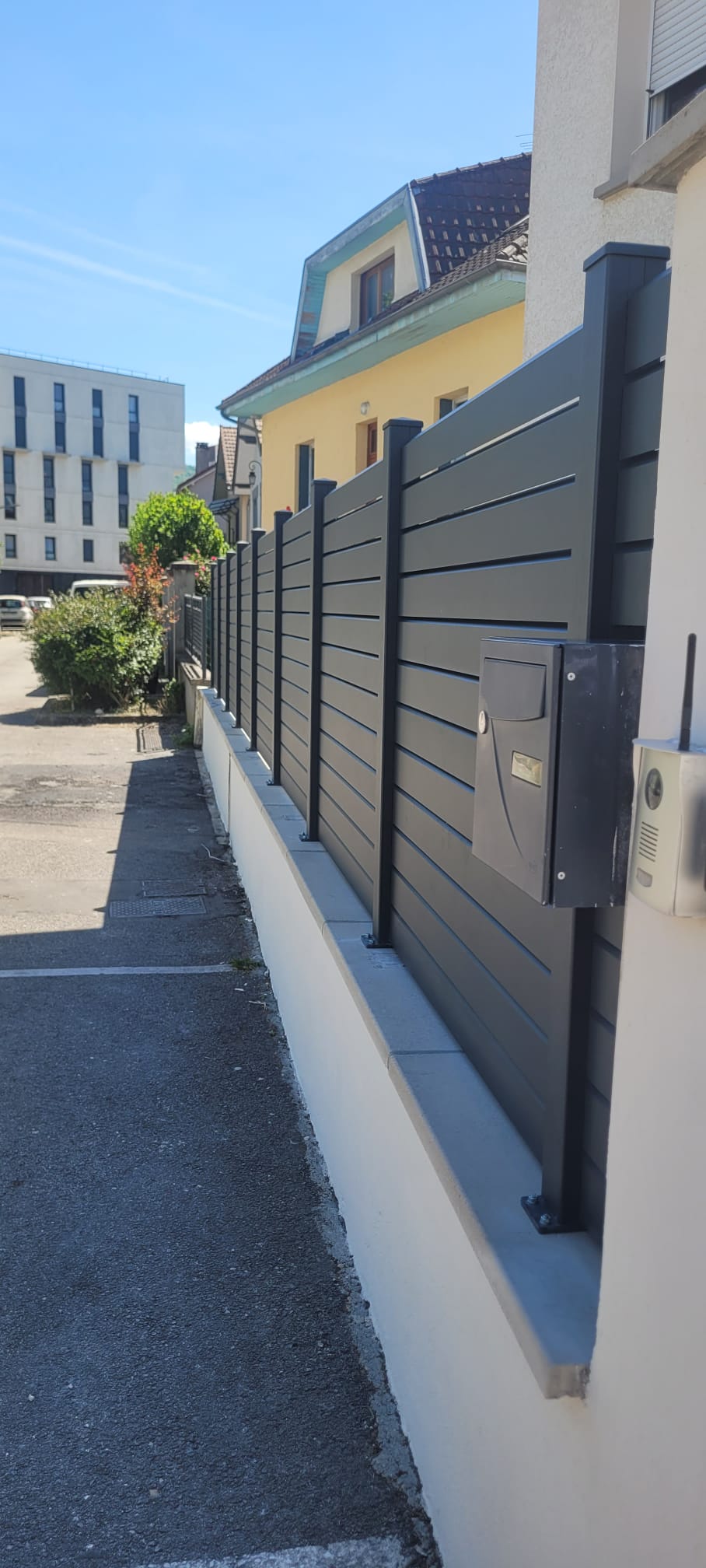 Entrée d'une maison moderne avec portillon en métal noir et chemin pavé sur herbe verte.