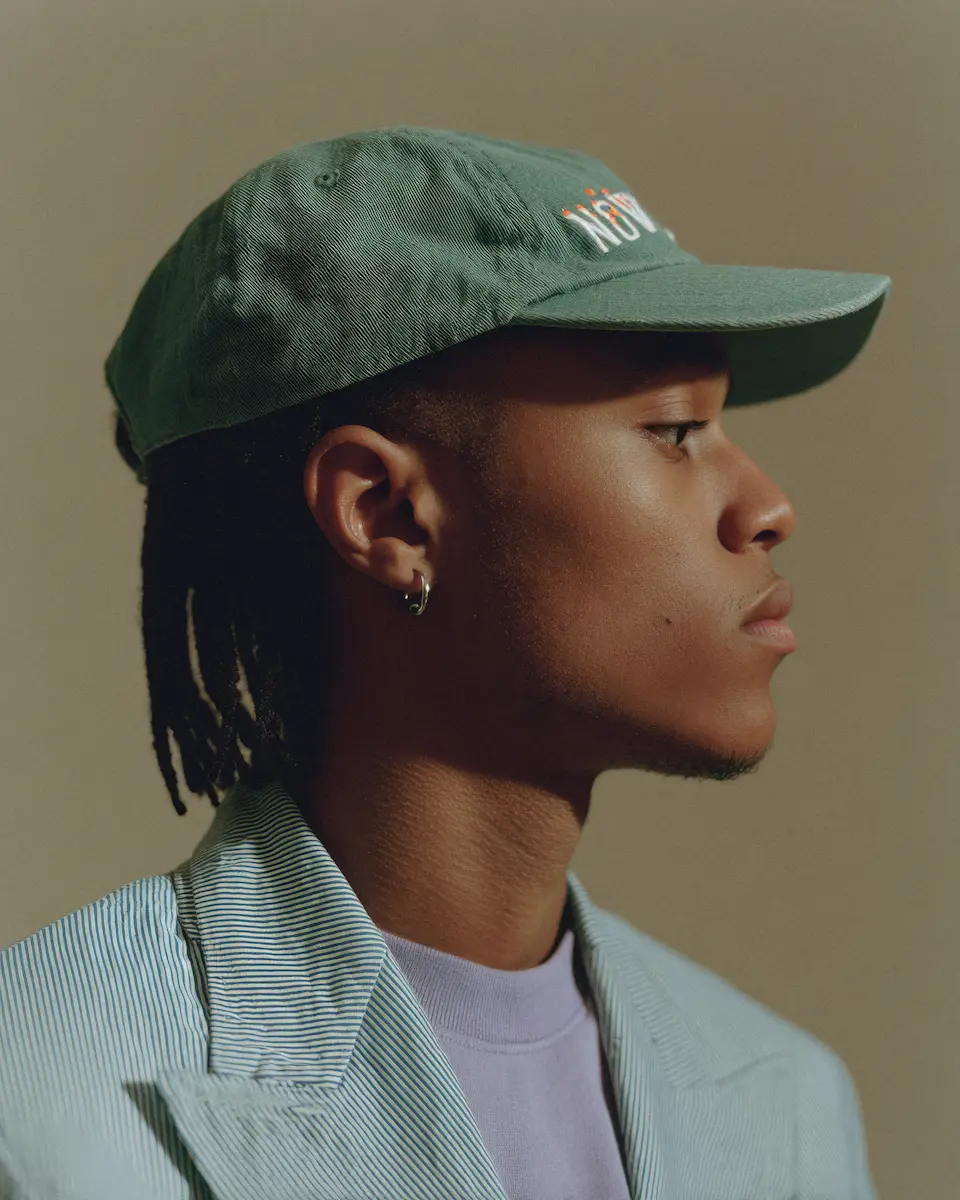 Profile of a person wearing a green cap, light striped jacket, and purple shirt looking to the right.