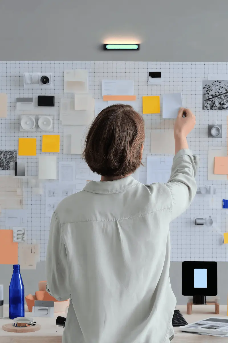 Person organizing sticky notes and papers on a grid wall in a creative workspace.