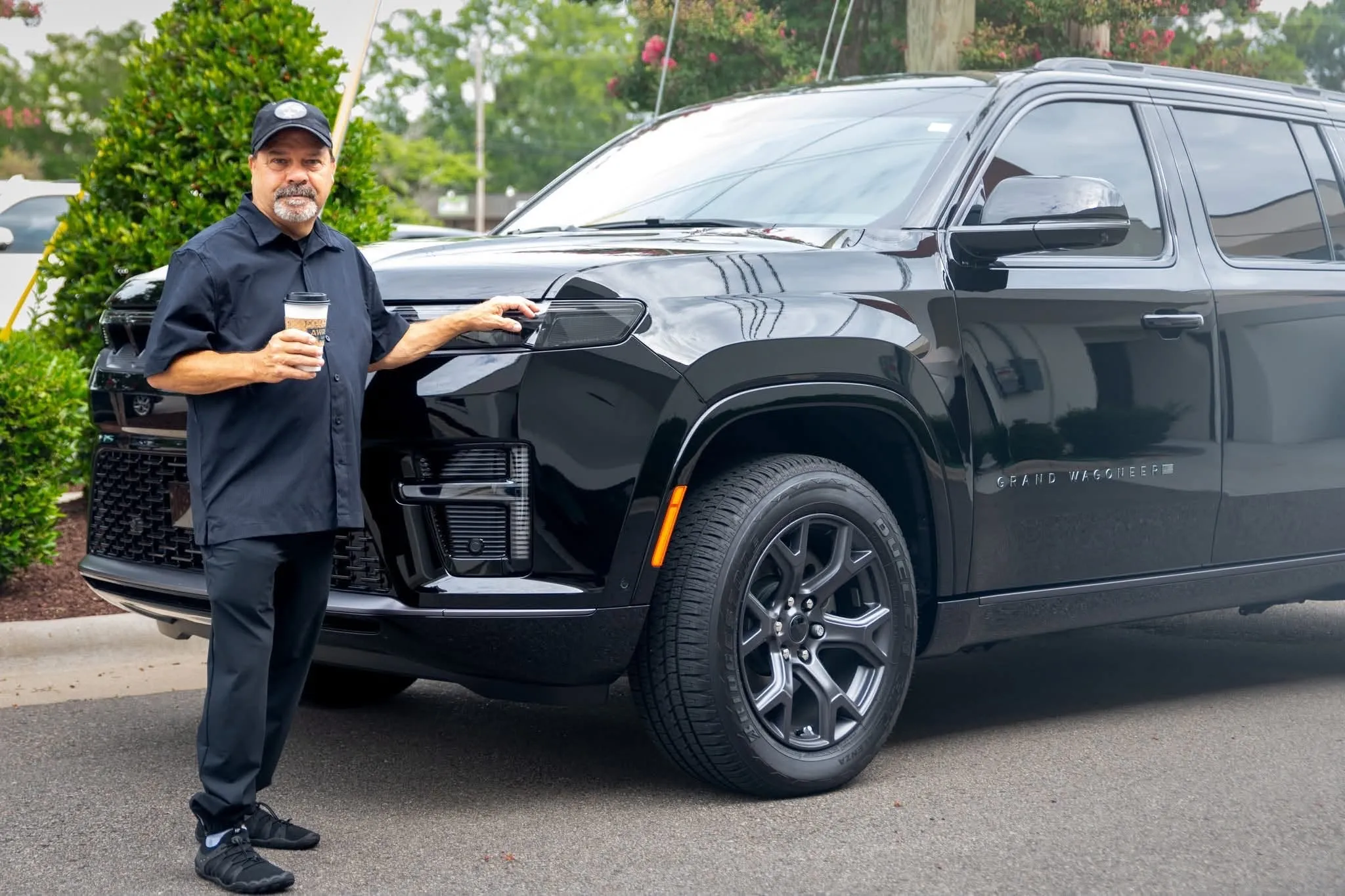 Richard Swindell, owner and driver of RM Executive Car Service, in black outfit holding coffee standing beside a shiny black Grand Wagoneer SUV outdoors.
