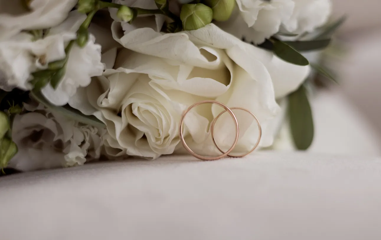 Two gold wedding rings resting against white roses and green leaves on a soft surface.