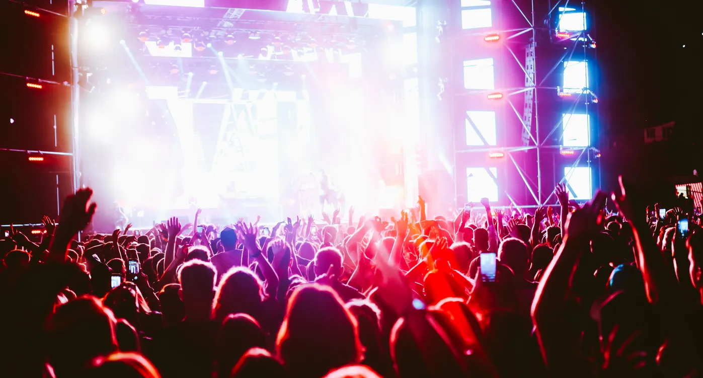 Large crowd with raised hands at a brightly lit outdoor music concert stage at night.