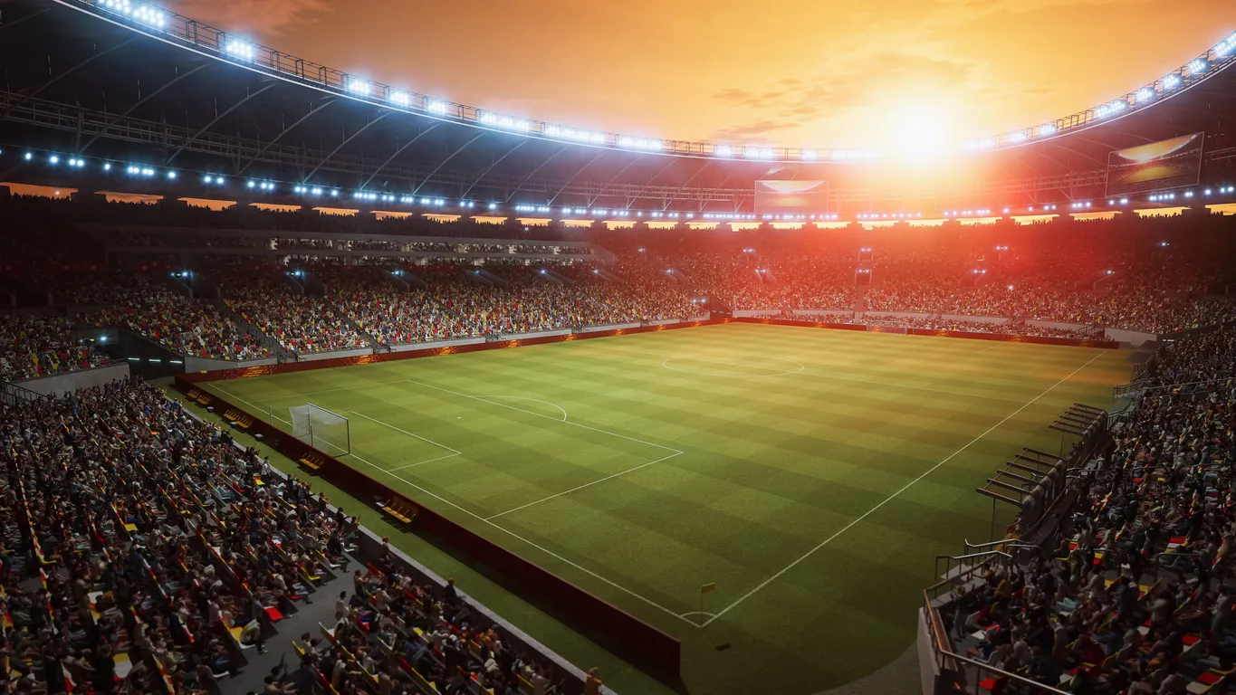 Wide view of a crowded soccer stadium with green field and sunset sky.