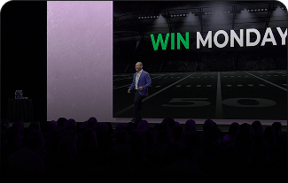A speaker in a suit stands on a stage in front of a screen displaying the words WIN MONDAY over a football field illustration with yard lines visible, addressing an audience in a dark auditorium.