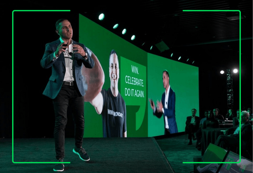 Man in a blazer and dark pants speaking on stage at a conference with a large screen behind him displaying another man wearing a 'WIN MONDAY' t-shirt and the text 'WIN. CELEBRATE. DO IT AGAIN.' Audience members are seated and watching the presentation.