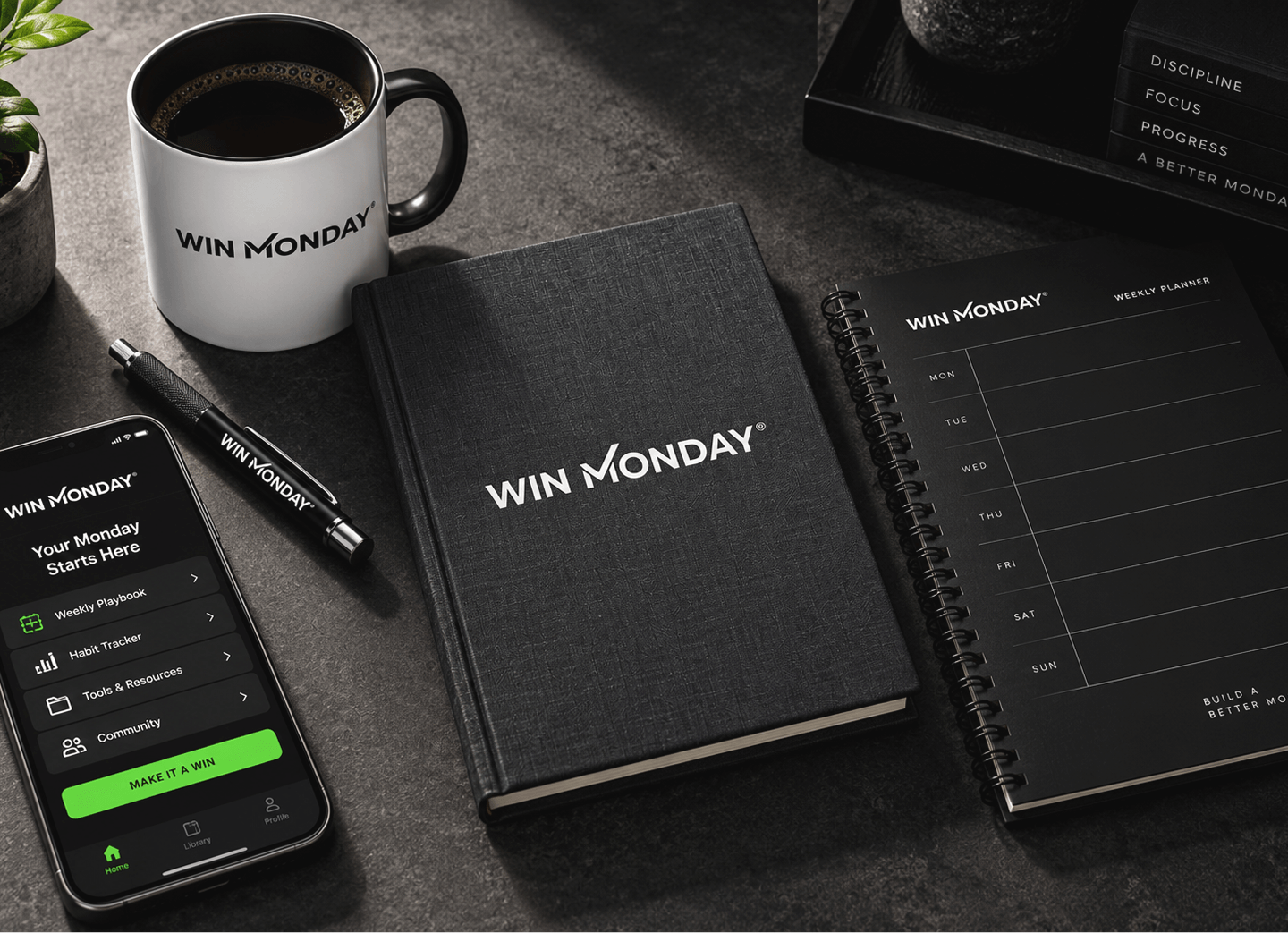 Desk setup with productivity tools branded Win Monday: a white coffee mug, a black pen, a closed planner, an open weekly planner, and a smartphone displaying the Win Monday app interface with sections like Weekly Playbook, Habit Tracker, Tools & Resources, and Community.