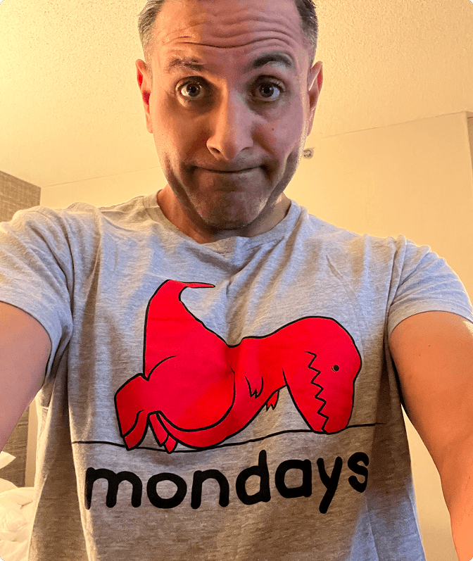 Man wearing a gray T-shirt with a red cartoon dinosaur lying down above the word 'mondays' in black letters, making a humorous face.