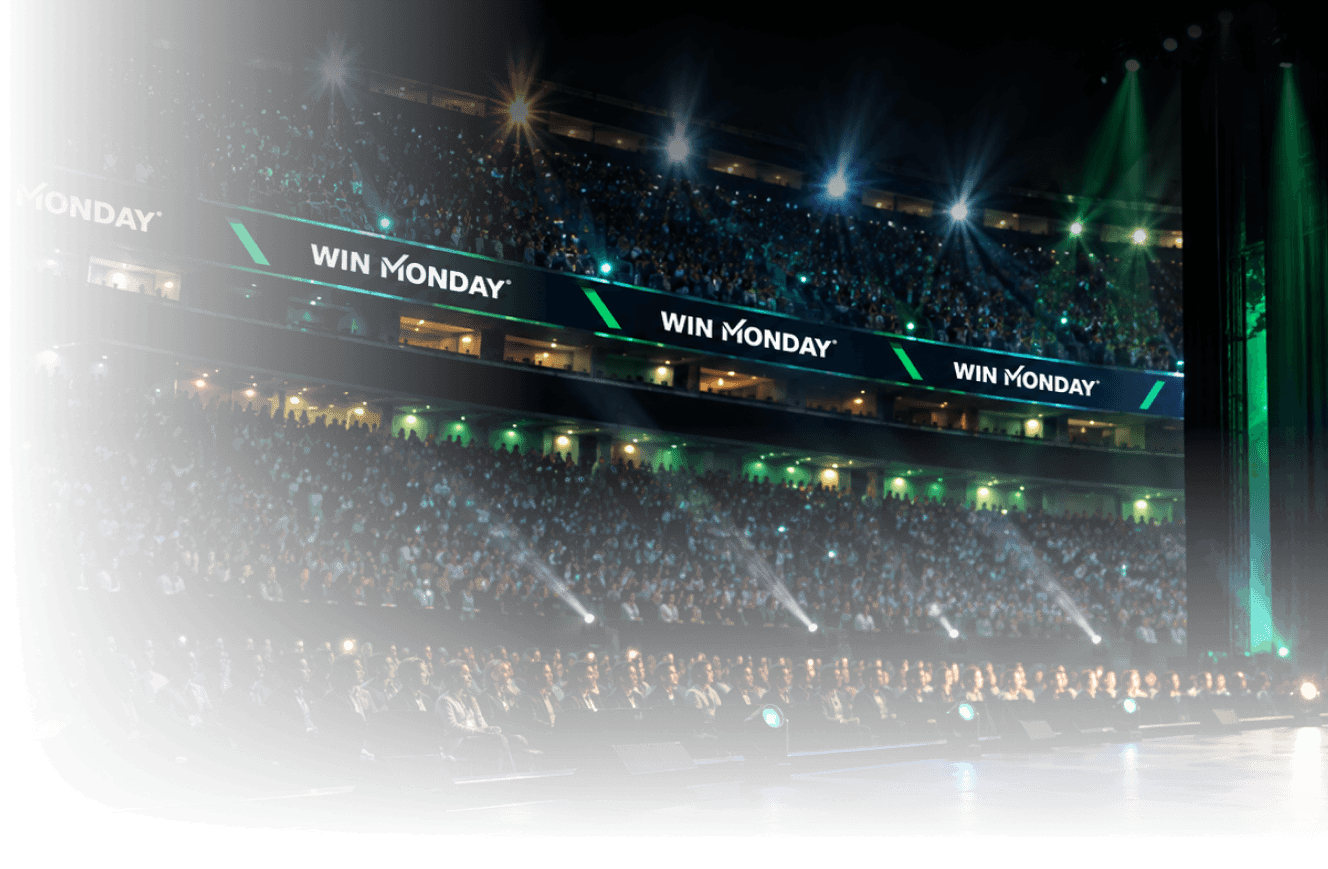 Large audience seated in a stadium or auditorium with bright stage lights and a digital banner displaying 'WIN MONDAY' repeatedly above the upper tier of seats.