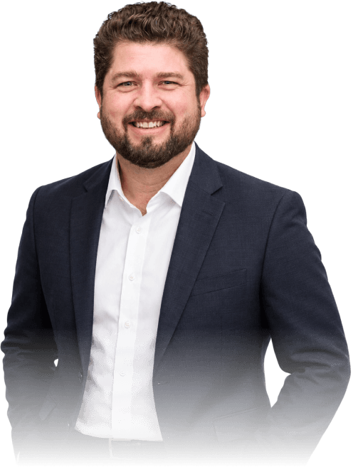 Smiling man with curly brown hair and beard wearing a navy blue suit jacket over a white button-up shirt, standing with hands in pockets.