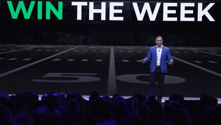 A man in a blue blazer and dark pants speaking on stage in front of an audience, with a large screen behind him displaying a football field and the words 'WIN THE WEEK' in bold letters.