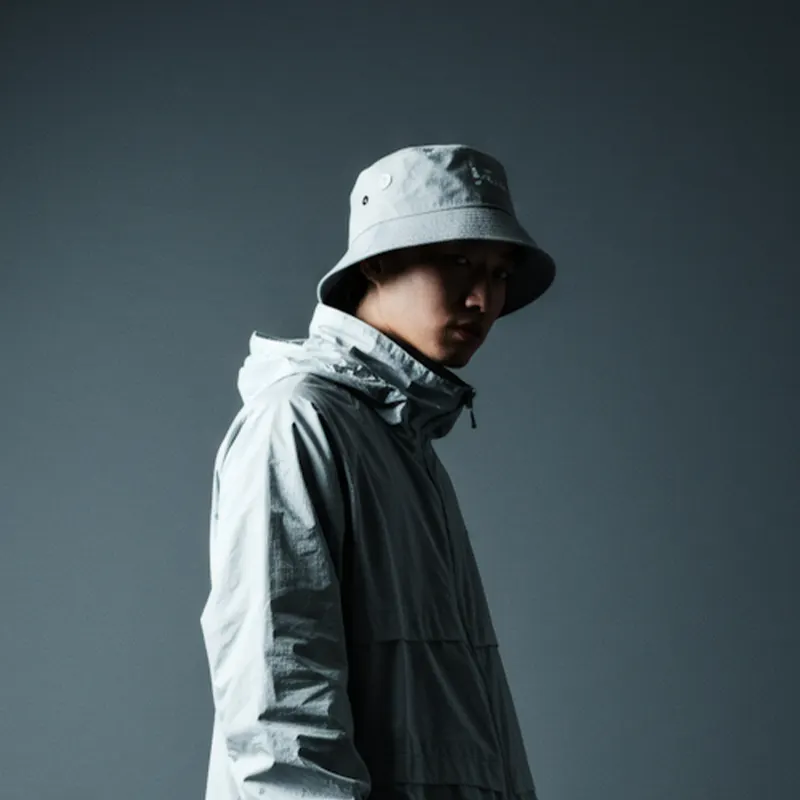 Person in a light gray bucket hat and matching hooded jacket against a dark gray background.