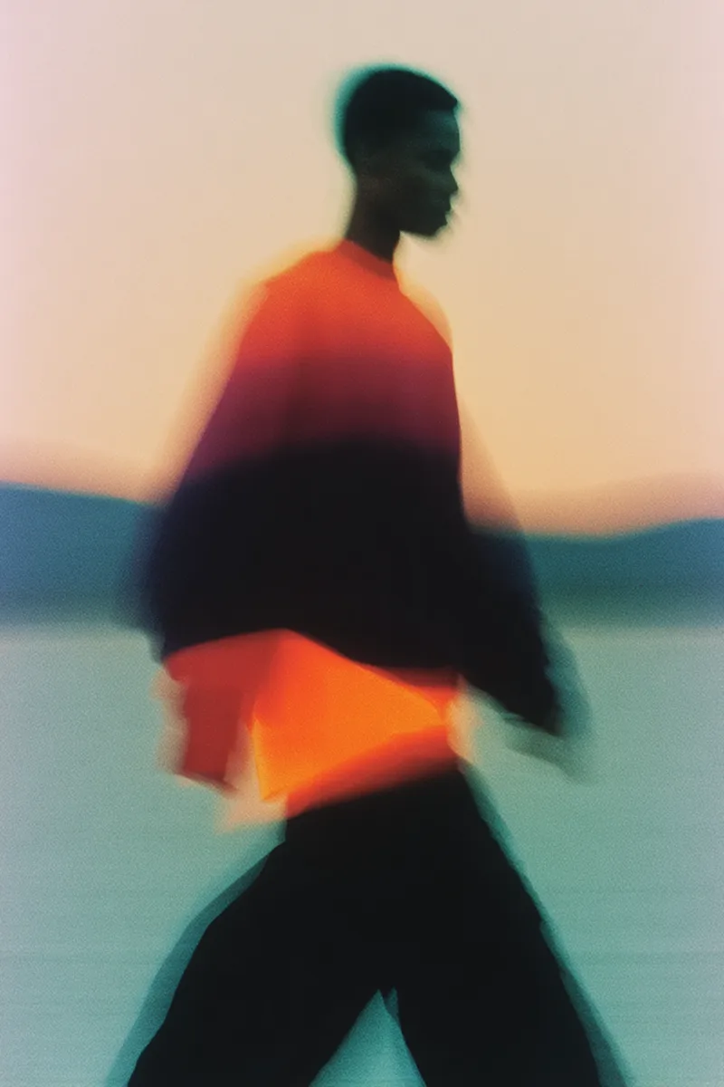 Blurred profile of a person walking, wearing a bright orange and black sweater against a pale sky.