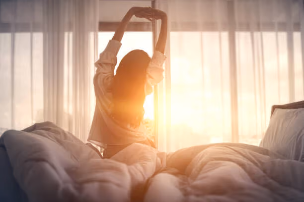 Person stretching while sitting on a bed facing a bright window with sheer curtains at sunrise.