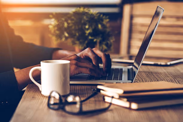 Hands typing on a laptop on a wooden desk with a white mug, notebooks, and glasses nearby.