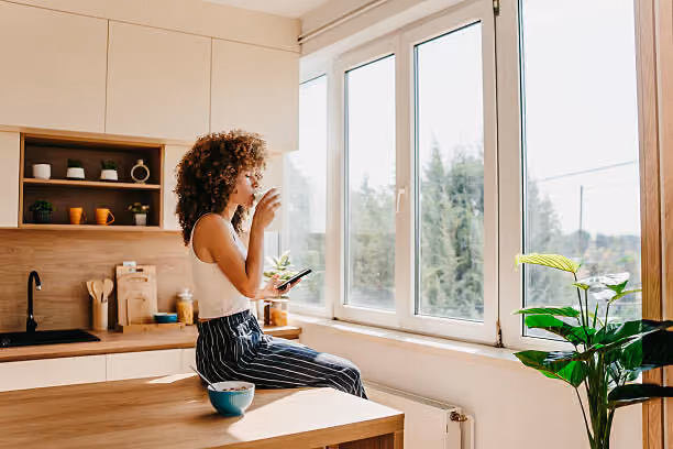 Woman in striped pants sitting on kitchen counter drinking coffee and holding phone near large windows.