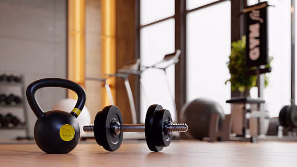 Black kettlebell and dumbbell resting on a gym floor with workout equipment blurred in the background.